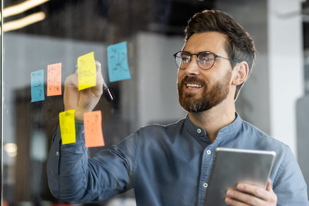 Businessman brainstorming and planning ideas on glass board using sticky notes while holding a...