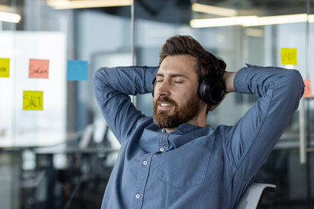 Close-up photo of young male office worker sitting in headphones at workplace and resting, eyes...