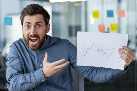 An angry young man sits in the office at the desk and emotionally shows documents with graphs to the...
