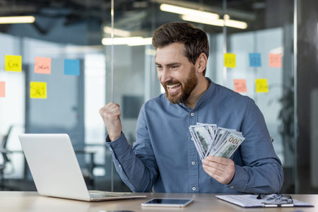 Young businessman looking excited holding money in hand while working on laptop in modern office....