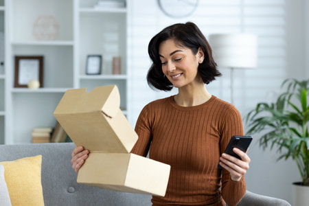 A smiling young woman is sitting on the sofa at home, holding a mobile phone and an open cardboard...