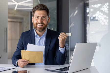 Joyful male executive with a brown envelope celebrates a business success at his desk in a modern...