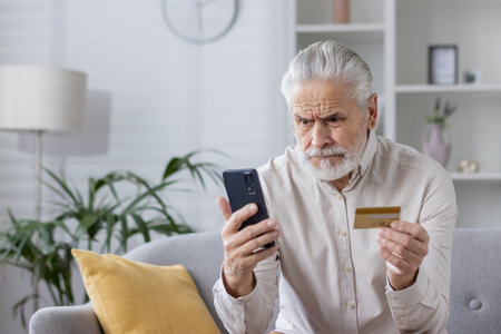Elderly man looking worried while checking his credit card information on his smartphone. Concept of...