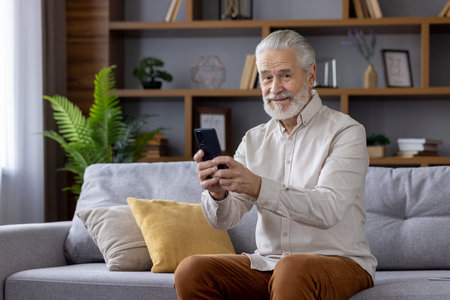 Portrait of smiling senior man grandfather in retirement sitting on sofa at home, holding mobile...