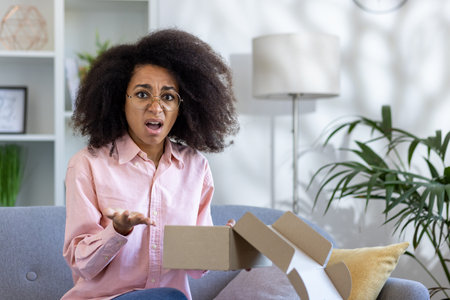 Woman expressing shock and confusion while opening package at home. Sitting on couch in living room...