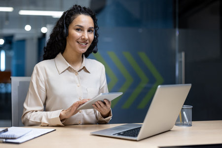 Smiling customer service representative using tablet and laptop at office desk wearing headset....