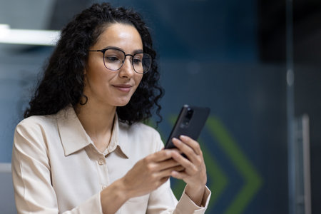 Confident professional woman wearing glasses using smartphone in modern office environment. Business casual attire and relaxed demeanor suggest productivity and focus in workplace.の写真素材