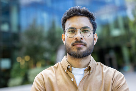 Confident man with glasses standing outdoors in front of modern building. Wearing casual shirt,...