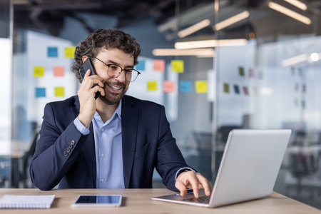 Businessman multitasking with laptop and phone in modern office. Professional in suit engaged in...
