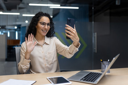 Confident businesswoman waving while engaging in video call. Modern office setting visualizing...