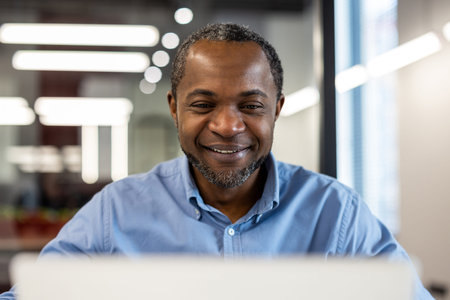 Smiling office worker wearing blue shirt working on laptop in modern office. Bright, professional...