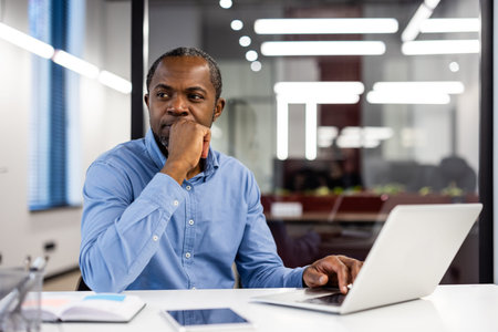 Pensive office worker in blue shirt thinking at desk with open laptop and documents. Modern office...