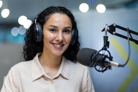 Smiling woman wearing headphones speaking into professional microphone in podcast studio. Confident...
