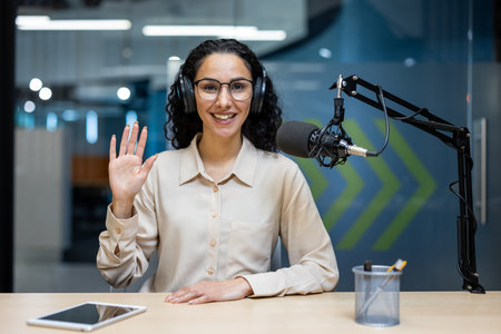 Smiling podcast host with headphones waving to camera in professional studio environment....