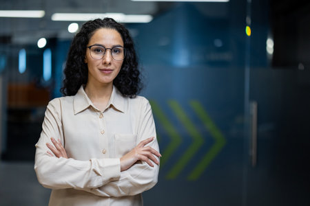Confident businesswoman with curly hair and glasses standing with arms crossed in modern office setting. Professional attire and serious expression convey leadership and determination.の写真素材