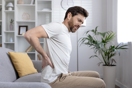 Man sitting on couch holding lower back, experiencing back pain, discomfort or injury. Concept of...