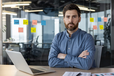 Business professional with arms crossed sitting at office desk with laptop and charts. Office...