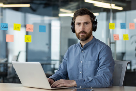 Confident professional man wearing headphones working on laptop in modern office. Colorful sticky...