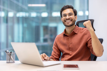 Smiling young professional celebrating success in modern office with laptop and phone on desk. Man expressing joy and achievement in business environment.の写真素材