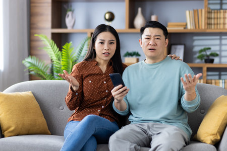 Asian couple sitting on sofa at home, both looking confused while holding smartphone. Concept of frustration, technology issue, misunderstanding, home lifestyle.の写真素材