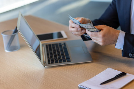 Business person counting dollar bills while using laptop and phone. Concept of finance, earnings, and digital work, with items including notebook, pen, and cup on desk.の写真素材