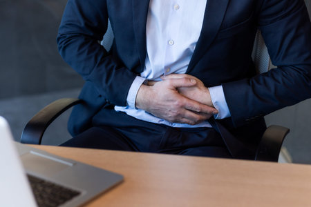 Businessman in formal suit sitting at office desk holding stomach in pain, indicating discomfort or...