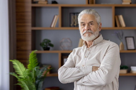 Portrait of a serious gray-haired old man with a beard standing in the office in a shirt, crossing...