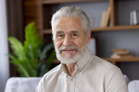 Portrait of a happy elderly man with grey hair and beard. He is sitting in a comfortable home living...