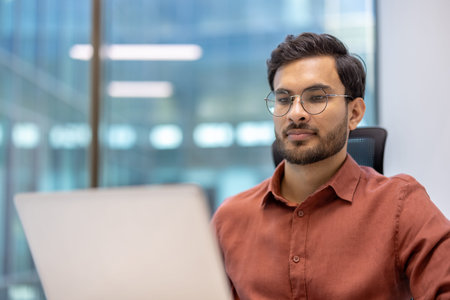 Young professional working on laptop in modern office setting. Focused employee wearing glasses...