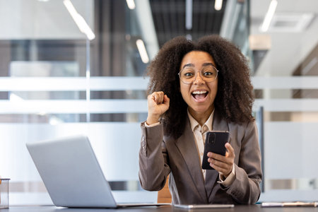 Young African American businesswoman excitedly using smartphone at workplace. Concept of success,...