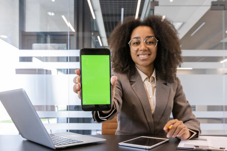Young African American businesswoman in professional attire presenting smartphone with green screen in office. Confident woman smiling and demonstrating technology with laptop and documents on deskの写真素材