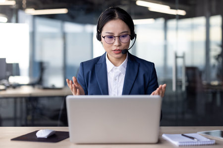Professional Asian businesswoman in formal attire using a headset for a video conference call in a...
