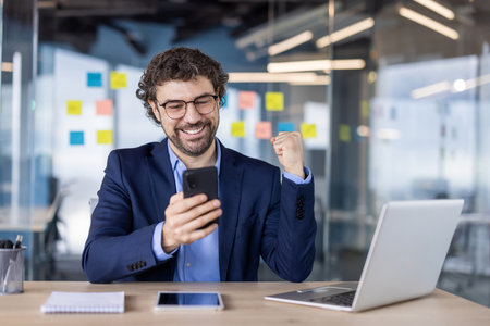 Happy businessman sitting at desk celebrating success while using phone in modern office. Smiling man in suit looking at smartphone with fist in air, laptop and tablet visible on desk.の写真素材