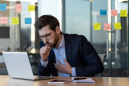 Businessman coughing and holding chest while working at laptop in office. Person experiencing health...