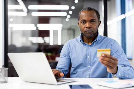 Serious businessman using laptop in office, holding credit card thoughtfully. Image shows focus and concentration in professional environment. Concepts include finance, technology, business tasks.の写真素材