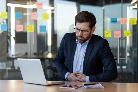 Business professional suffers from stomach pain while working at desk. Man in formal attire shows...