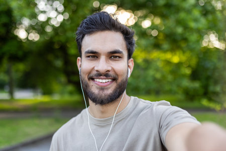 Young man smiling and taking selfie with phone in outdoor park. Enjoying nature and listening to music with earphones. Casual wear and cheerful expressions.の写真素材