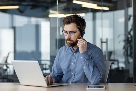 A young serious man works in the office on a laptop, wears a headset and talks on a video call,...