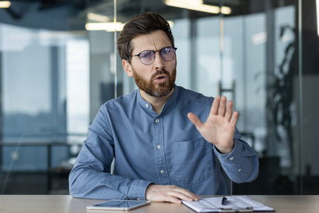 A young serious man in glasses and a blue shirt is sitting in the office at the desk and talking...