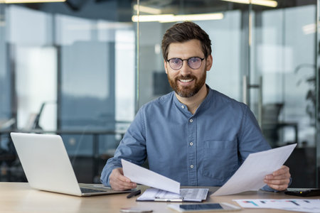 Portrait of a smiling and successful young businessman sitting in the office at the table, holding...