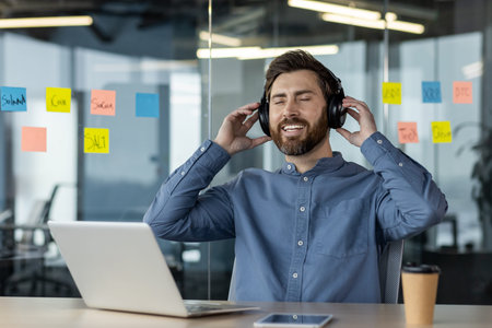 A young smiling man is sitting in the office at the work desk wearing headphones, closed his eyes...