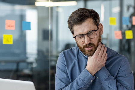Close-up photo of a young man working in the office, sitting at the desk and holding his cheek with...