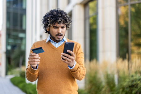 Concerned man using phone outdoors, holding credit card, experiencing trouble online banking or...