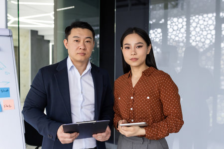 Asian business professionals stand confidently in office setting, holding digital tablets. They exude professionalism and determination, representing work, teamwork, success, and technologyの写真素材