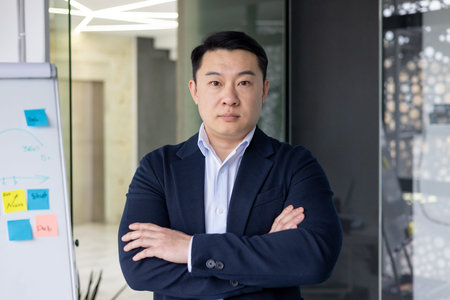 Confident asian businessman standing with arms crossed in modern office environment. Whiteboard with...