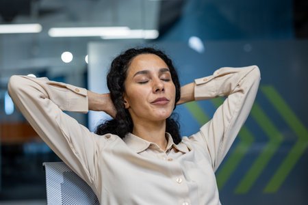 Confident businesswoman in office takes a moment to relax with closed eyes and hands behind head. Wearing a smart white shirt, she embodies calm and tranquility during workplace break.の写真素材