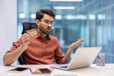 Man sitting at desk holding credit card while using laptop, showing frustration and confusion with...