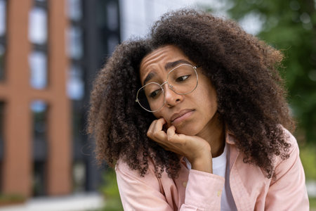 African American woman with curly hair and glasses appears thoughtful and contemplative. Wearing...
