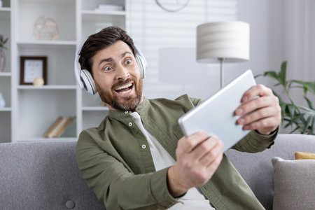 Joyful man with headphones holding tablet intensely focused on video games. Expresses excitement and engagement in modern gaming experience using phone and technology.の写真素材