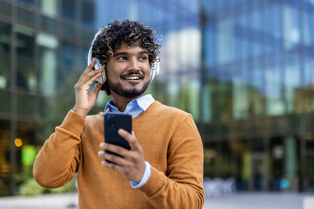 Young man wearing headphones holds smartphone, enjoying music with relaxed expression. Captures modern lifestyle of urban professional connecting digitally during work breakの写真素材